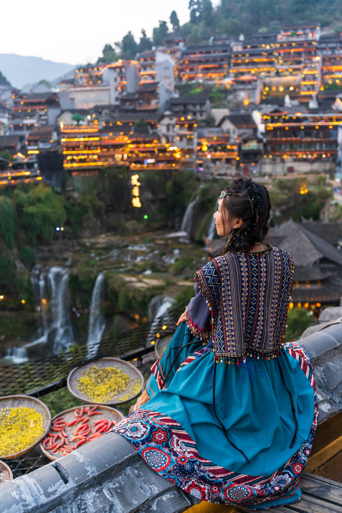 Una mujer de espaldas, con un traje étnico de colores, está sentada en un mirador contemplando una ciudad iluminada en una colina al anochecer, con una cascada al lado