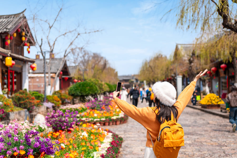 mujer turista usando sueter y mochola amarilla mientras pasea en un calle con flores